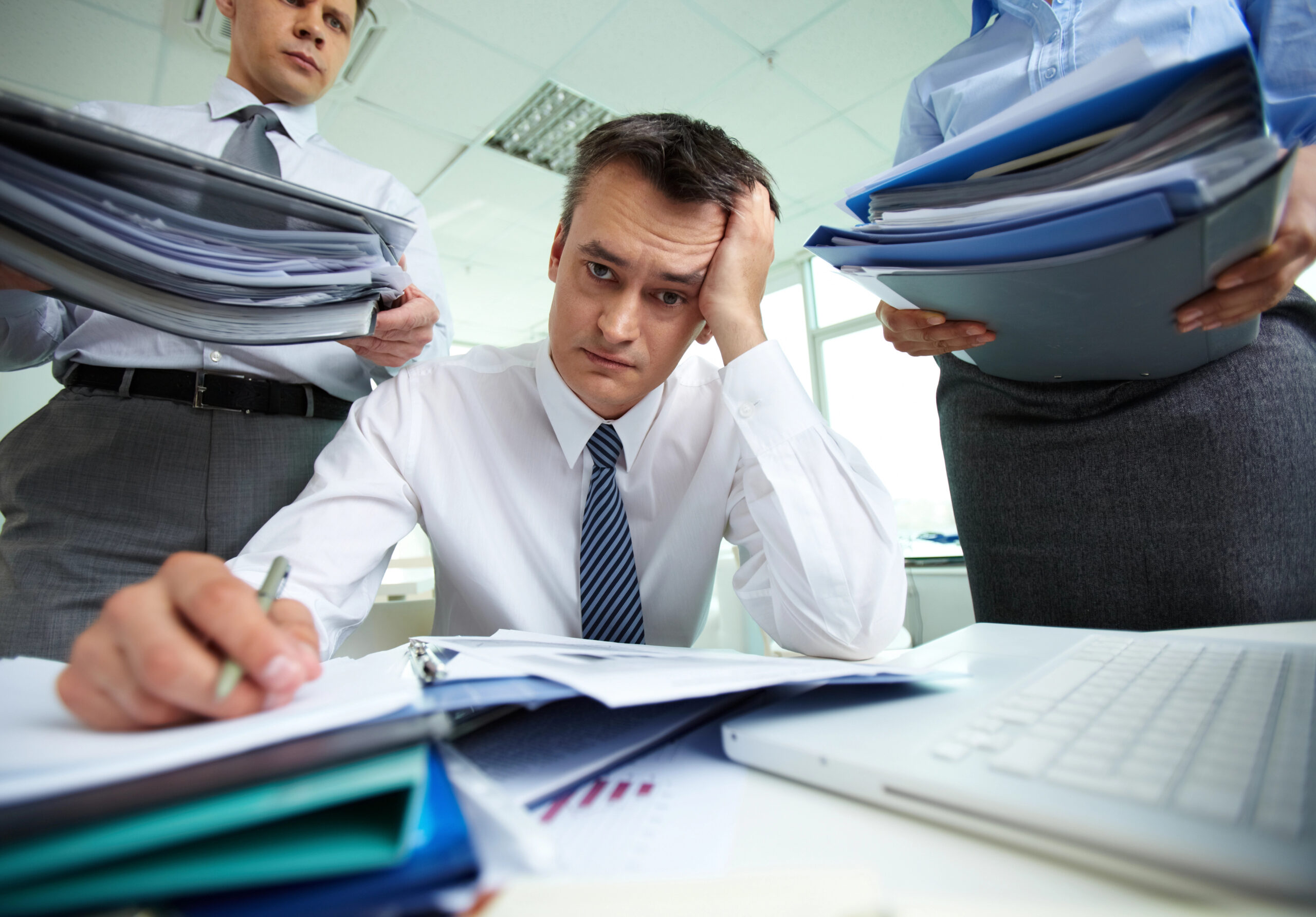 Businessman looking stressed leaning over a stack of papers with coworkers standing behind him